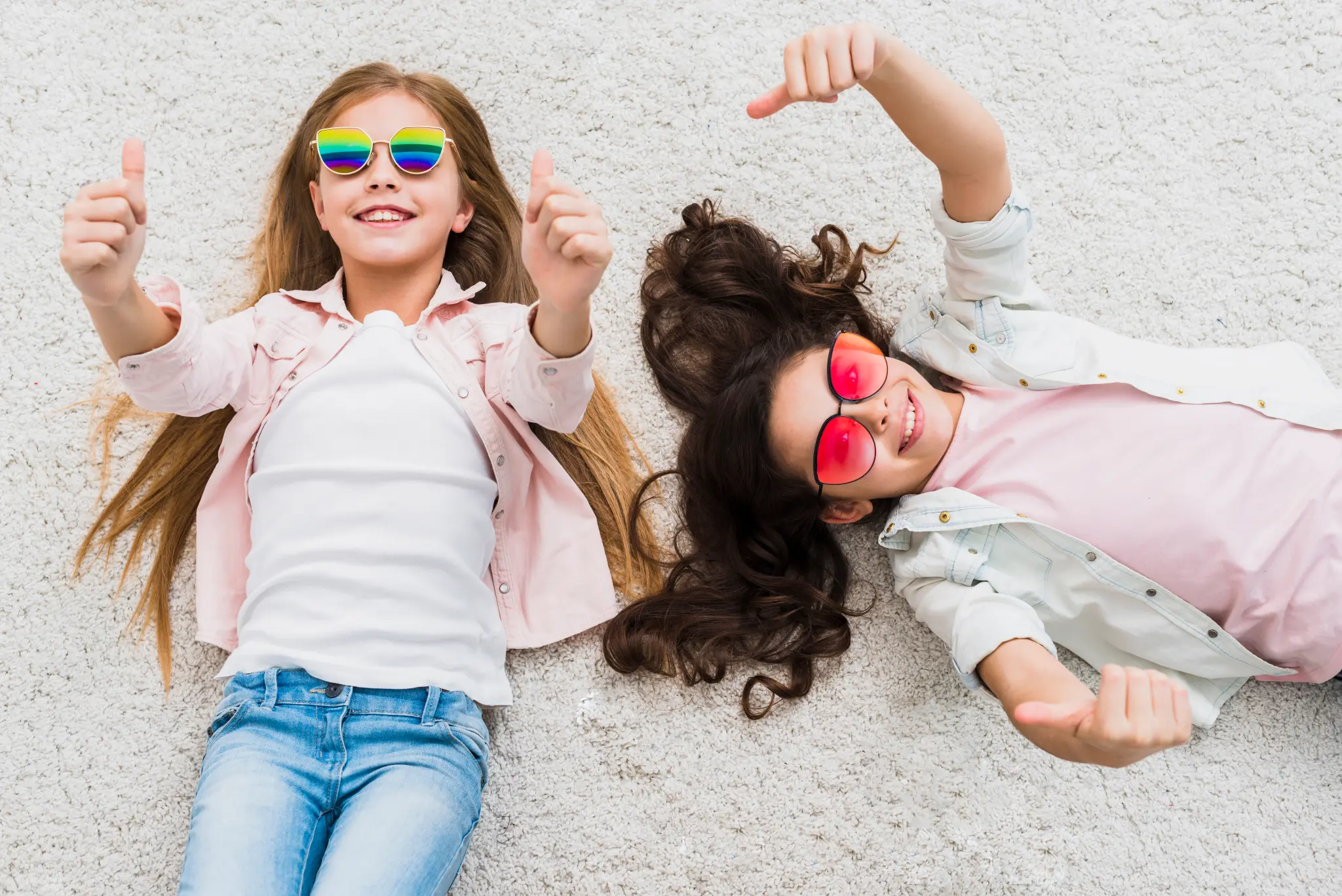 Two young girls lying on a carpet wearing sunglasses and giving thumbs up